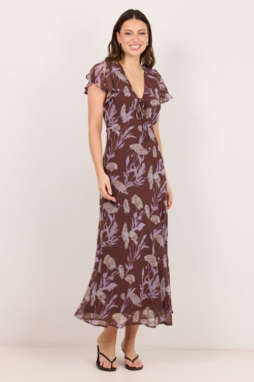 A woman modeling a brown maxi dress with a purple floral pattern, ruffled short sleeves, and a V-neckline, standing against a plain off-white studio background.