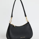 Black pebble-textured leather shoulder bag with gold hardware, featuring a curved silhouette, double adjustable straps and top zip closure. Stylish small black handbag from Among the Brave, ideal for everyday wear.