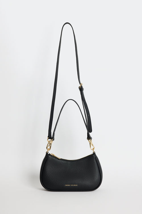 Black pebble-textured leather shoulder bag with gold hardware, featuring a curved silhouette, double adjustable straps and top zip closure. Stylish small black handbag from Among the Brave, ideal for everyday wear.