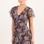 A woman modeling a short brown mini dress with a white paisley pattern, flutter sleeves, and a V-neckline, photographed against a plain off-white studio background.