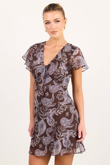 A woman modeling a short brown mini dress with a white paisley pattern, flutter sleeves, and a V-neckline, photographed against a plain off-white studio background.