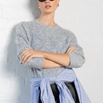 Person wearing a gray sweater, light blue shirt, black skirt, and black shoes against a white wall.