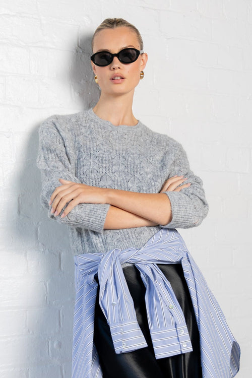 Person wearing a gray sweater, light blue shirt, black skirt, and black shoes against a white wall.