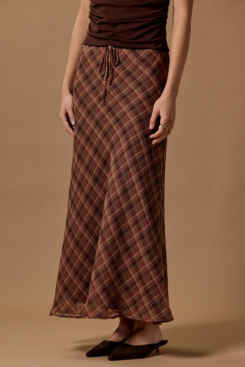A model wearing a brown and red plaid maxi skirt with a drawstring waist, paired with a dark brown top and black pointed flats, against a solid beige studio background.