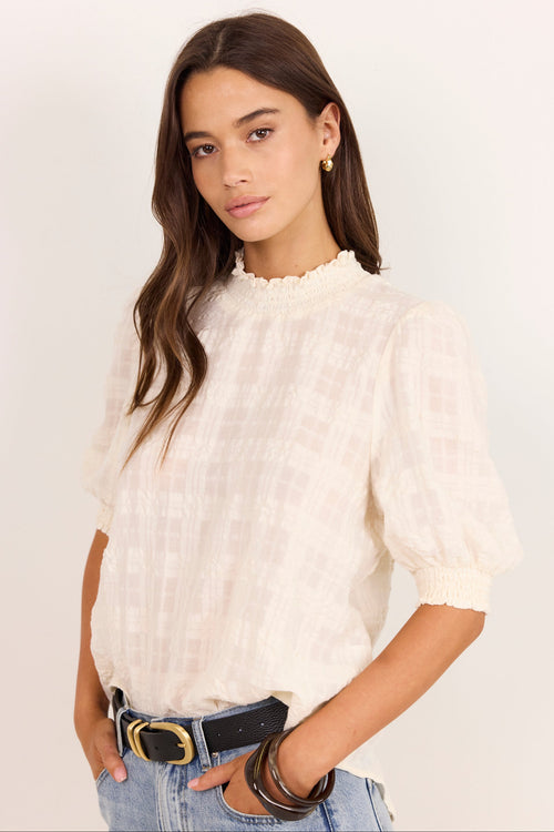 A woman modeling a short-sleeve white textured blouse with a ruffled mock neck and puffed sleeves, paired with blue jeans and a black belt, against a plain white studio background.