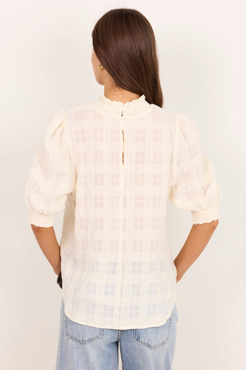 A woman viewed from the back wearing a cream-colored textured blouse with puff sleeves and a high ruffled collar, paired with light blue jeans, against a plain white background.