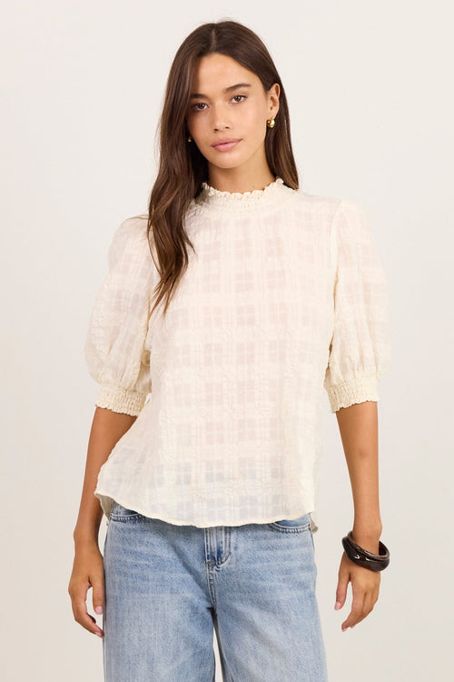 A woman modeling a cream-colored textured puff-sleeve blouse with a subtle grid pattern, paired with light-wash wide-leg jeans and black sandals, standing against a plain off-white studio background.