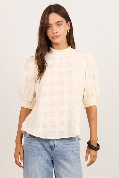 A woman modeling a cream-colored textured puff-sleeve blouse with a subtle grid pattern, paired with light-wash wide-leg jeans and black sandals, standing against a plain off-white studio background.