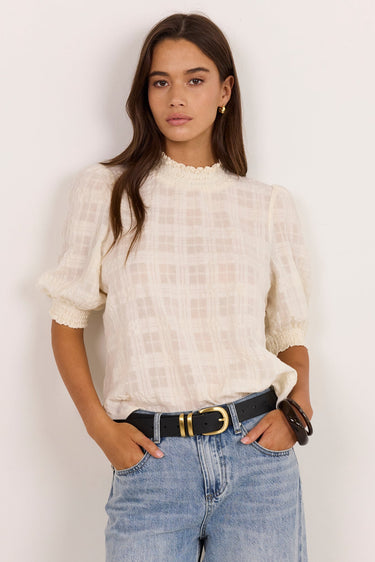 A woman modeling a cream-colored textured puff-sleeve blouse with a subtle plaid pattern, tucked into light-wash blue jeans with a black belt, standing against a plain off-white wall.
