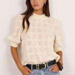 A woman modeling a cream-colored textured short-sleeve blouse with a ruffled high neck, paired with light-wash blue jeans and a black belt, standing against a plain off-white background.