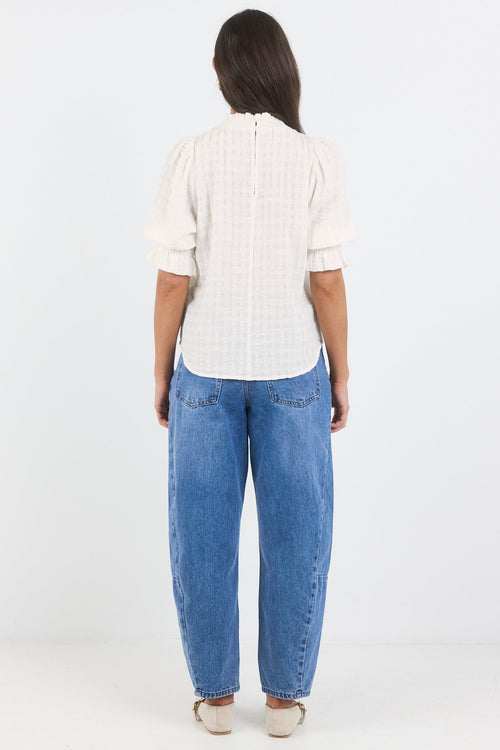 A woman viewed from the back wearing a white short-sleeve checkered blouse and blue jeans, standing against a plain white studio background.