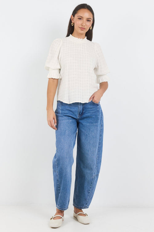 A woman modeling a white textured short-sleeve blouse with puffed shoulders and blue wide-leg jeans, standing against a plain white studio background.