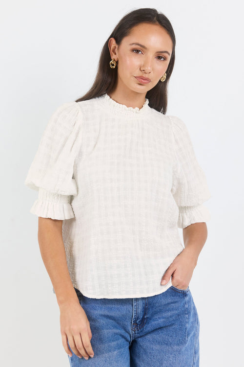 A woman modeling a white short-sleeve blouse with a subtle grid pattern, ruffled collar, and voluminous flutter sleeves, paired with blue jeans against a plain white studio background.