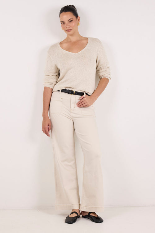 A woman modeling a light beige knit long-sleeve top and matching wide-leg trousers, styled with a black belt and black flat shoes, against a plain white studio background.