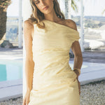 Woman wears a soft yellow one-shoulder mini dress with side ruching that creates a flattering, figure-skimming shape. The look is sleek, minimal, and slightly playful.