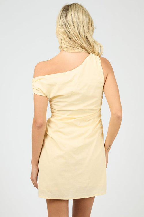 Woman wears a soft yellow one-shoulder mini dress with side ruching that creates a flattering, figure-skimming shape. The look is sleek, minimal, and slightly playful.