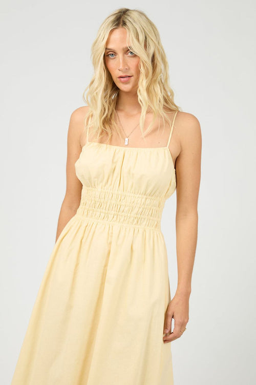 Woman wears a lemon yellow maxi dress with a smocked bodice and thin spaghetti straps. The dress flows gently from the waist, styled with strappy heels for a light, airy feel.
