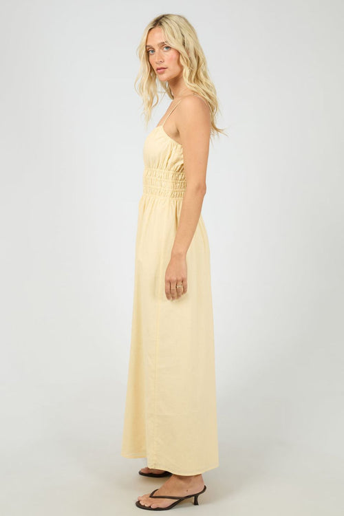 Woman wears a lemon yellow maxi dress with a smocked bodice and thin spaghetti straps. The dress flows gently from the waist, styled with strappy heels for a light, airy feel.