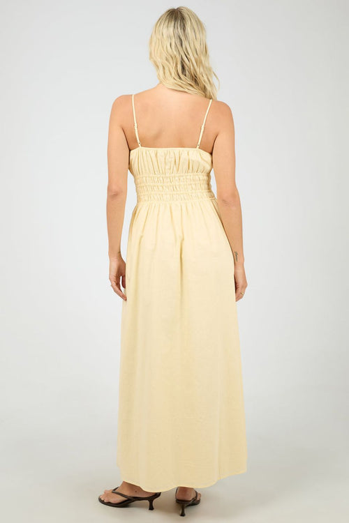Woman wears a lemon yellow maxi dress with a smocked bodice and thin spaghetti straps. The dress flows gently from the waist, styled with strappy heels for a light, airy feel.