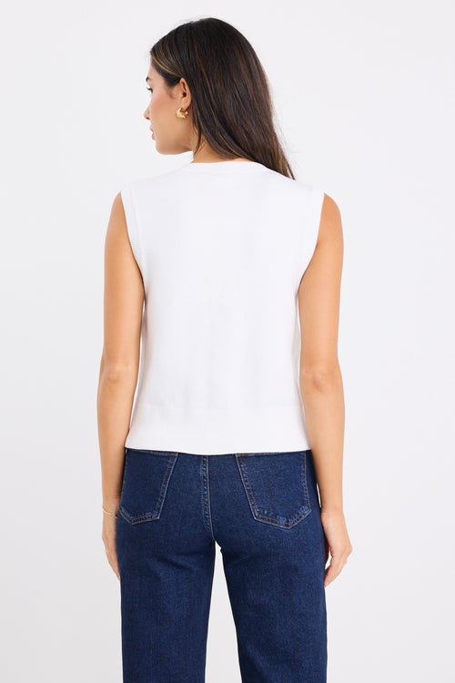 Woman wearing a sleeveless cream vest with button-down front and crew neck, paired with dark denim jeans.