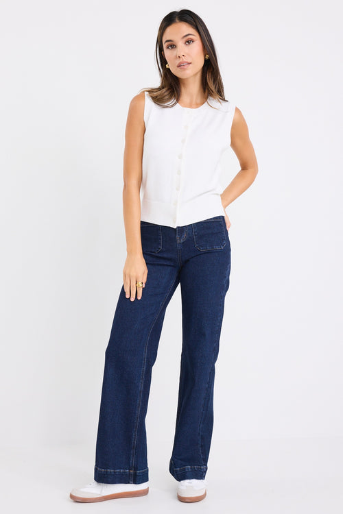 Woman wearing a sleeveless cream vest with button-down front and crew neck, paired with dark denim jeans.