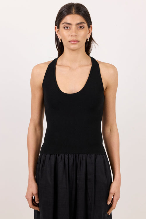 A woman modeling a black sleeveless scoop-neck top paired with a black pleated skirt, standing against a plain off-white studio background.