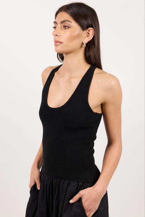 A woman modeling a black ribbed knit tank top with a wide scoop neckline, paired with a black skirt, against a plain off-white studio background.