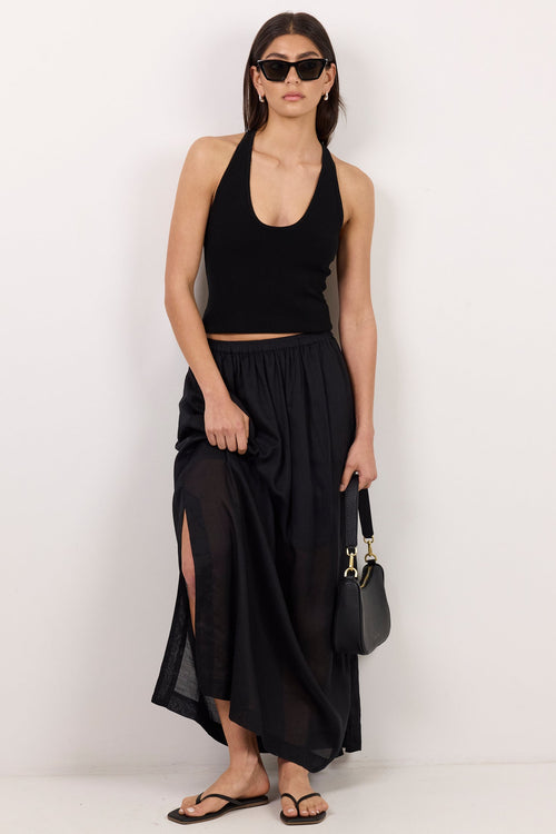 A model stands against a plain white studio wall wearing a black sleeveless crop top, a long black skirt with a side slit, black sandals, and holding a small black handbag.