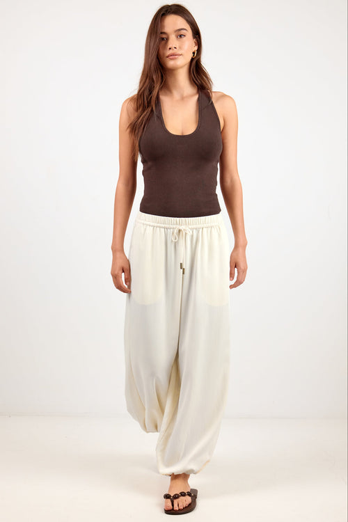 A woman modeling loose, draped cream-colored pants with a drawstring waist, paired with a dark brown tank top and brown sandals, standing against a plain white studio background.