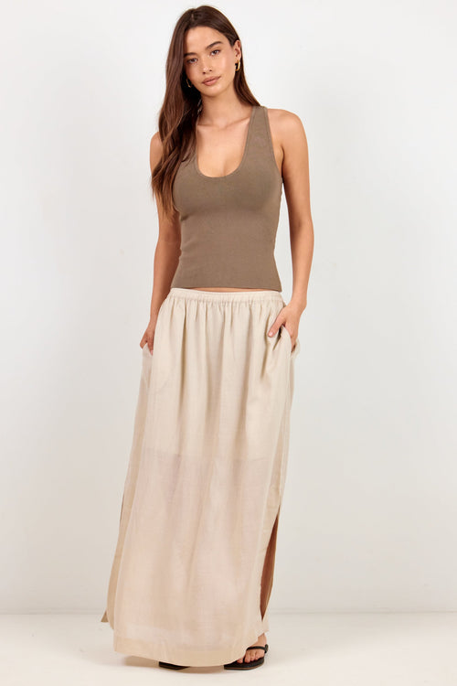 A woman modeling a two-piece outfit consisting of a taupe brown ribbed tank top and a long, flowing light beige skirt with side slits, set against a plain white studio background.