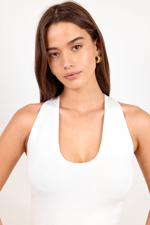 A woman wearing a white halter-neck tank top and small gold hoop earrings, photographed against a clean white studio background.