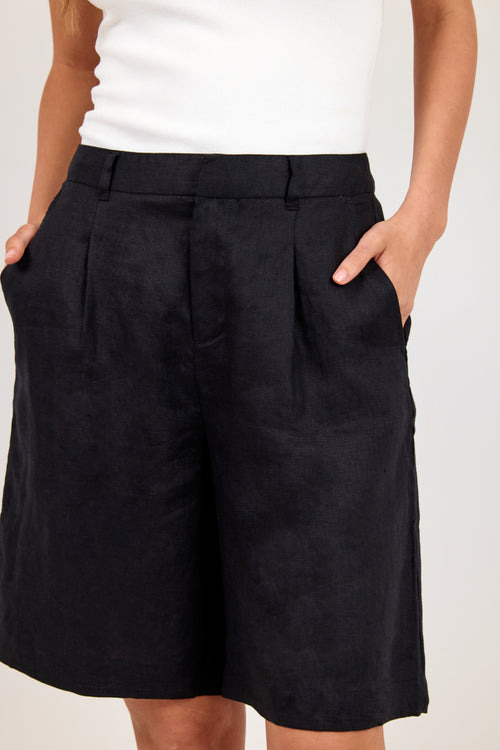 A close-up of a model wearing black pleated linen shorts with side pockets, paired with a white top, against a plain light background.