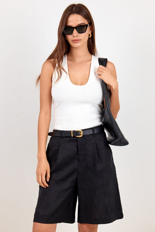 A woman modeling a white ribbed tank top, black pleated shorts, a black belt with a gold buckle, and black sunglasses, holding a black shoulder bag against a plain light background.