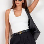 A woman modeling a white ribbed tank top, black pleated shorts with a black belt, black sunglasses, and carrying a black shoulder bag against a plain white background.