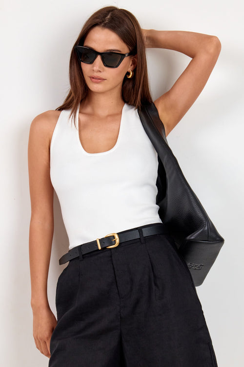 A woman modeling a white ribbed tank top, black pleated shorts with a black belt, black sunglasses, and carrying a black shoulder bag against a plain white background.