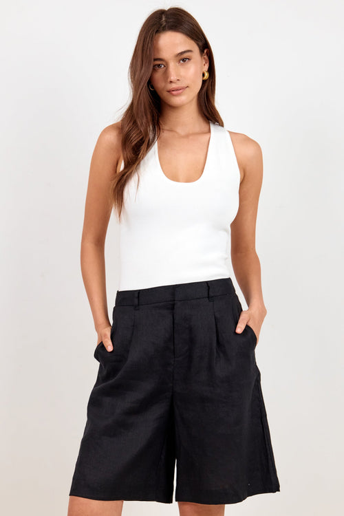 A woman modeling a white ribbed tank top and black wide-leg shorts against a plain light gray studio background.