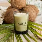 A lit candle in a clear glass jar with the brand name 'OASIS' printed on the front, styled on a table with palm leaves, coconuts, and glassware.