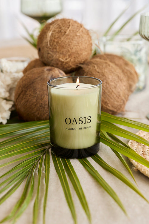 A lit candle in a clear glass jar with the brand name 'OASIS' printed on the front, styled on a table with palm leaves, coconuts, and glassware.
