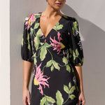 A woman modeling a black midi dress with a vibrant pink, green, and yellow floral print, standing against a neutral gray wall.