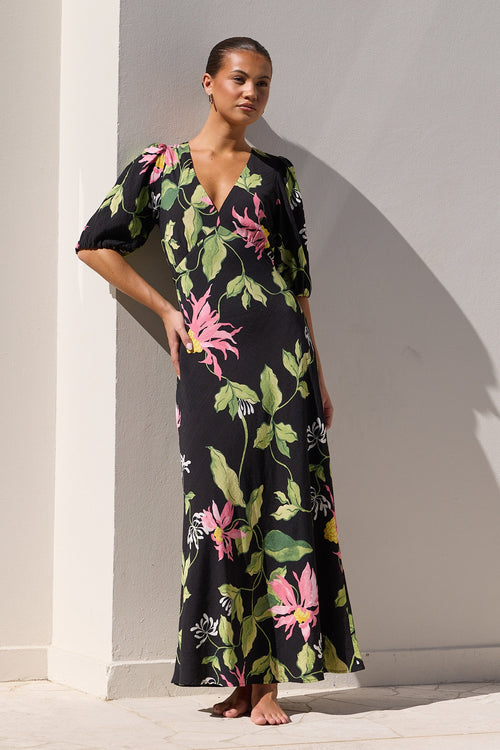 A woman stands barefoot at the edge of a swimming pool wearing a black maxi dress with a vibrant pink and green tropical floral print, posing against a white wall.