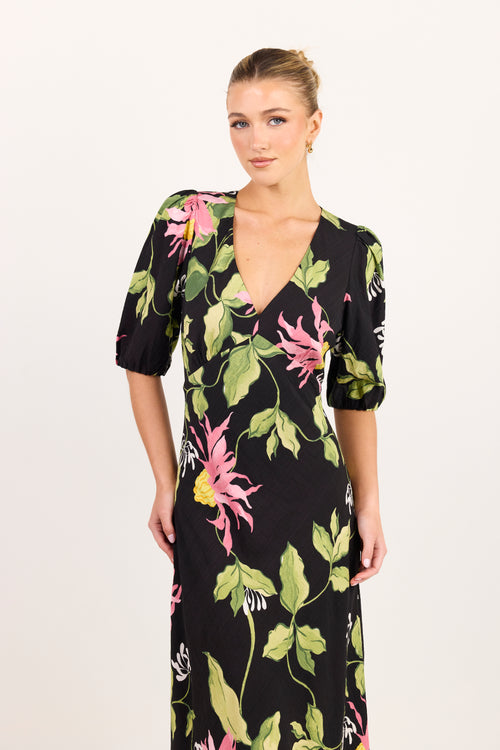 A woman modeling a black midi dress with a vibrant pink and green floral print, featuring puff sleeves and a V-neckline, set against a plain light background.