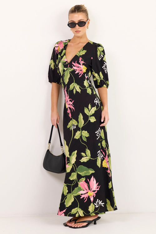 A woman modeling a black maxi dress with a vibrant pink and green floral print, styled with a black handbag and sunglasses against a plain white studio background.