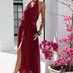 A woman models a sleeveless, halter-neck, midi-length red maxi dress with a high side slit and a matching belt with a circular buckle. She is posing outdoors on a sunny patio with white columns and potted pink flowers.