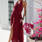 A woman models a sleeveless, deep burgundy halter-neck maxi dress with a wrap-style bodice and a matching tie belt at the waist. She is standing outdoors in front of a white building, with a large planter containing bright pink flowers on the right side of the frame.