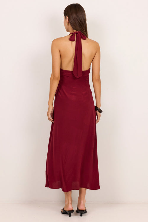 A woman viewed from the back wearing a sleeveless halter-neck maxi dress in deep burgundy, paired with black pointed-toe flats, set against a plain off-white studio background.