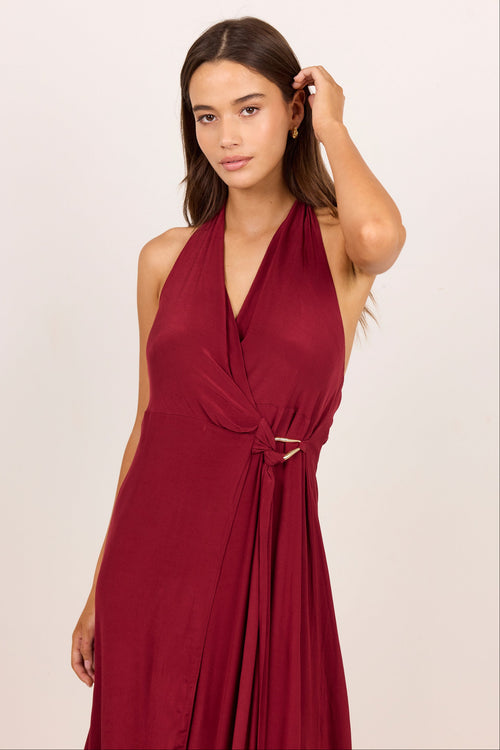 A woman modeling a sleeveless halter-neck maxi dress in deep burgundy with a wrap-style V-neck and a tie detail at the waist, set against a plain light beige studio background.
