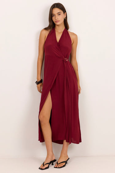 A woman modeling a sleeveless halter-neck midi dress in deep burgundy with a wrap-style front and high side slit, standing against a plain off-white studio background.