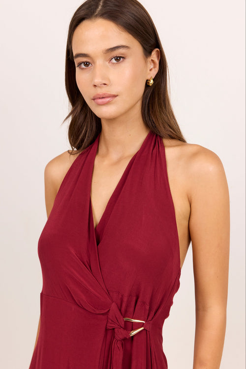 A woman wearing a sleeveless halter-neck wrap dress in deep burgundy with a twisted waist tie and a small gold buckle detail, photographed against a plain light beige studio background.