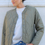 Woman wearing a green quilted jacket and blue jeans against a white wall