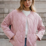 Woman wearing a pink quilted jacket and blue jeans against a brick wall.